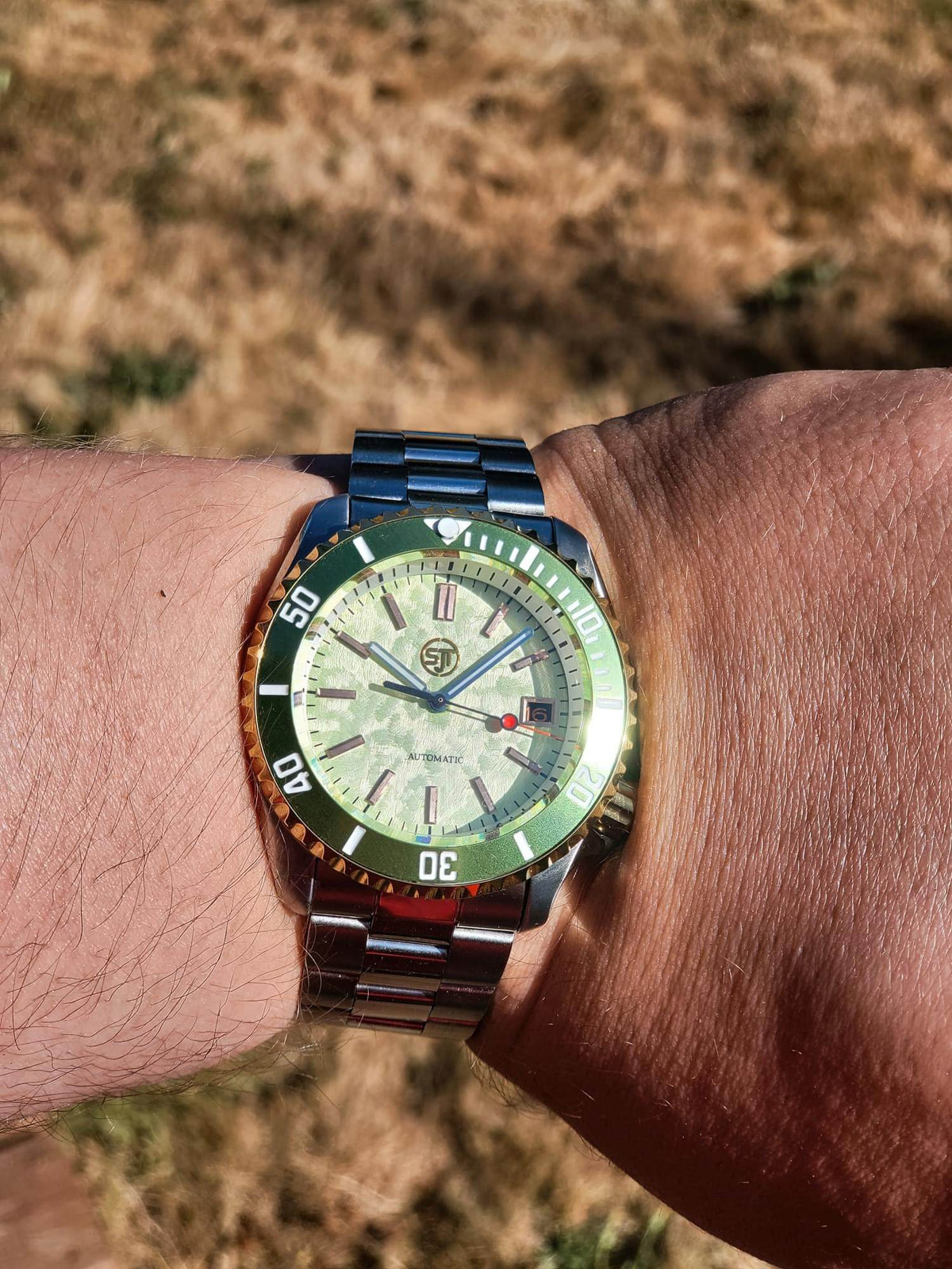 The Coconut SJT Original wristwatch with a green bezel, silver-toned metal bracelet, and a cream-colored dial, shown worn on a person's wrist against a blurred outdoor background.