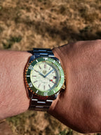 A close-up of a wrist wearing The Coconut SJT Original wristwatch with a green bezel, a cream dial, and a matching green and steel bracelet, set against a blurred outdoor background of dry grass.