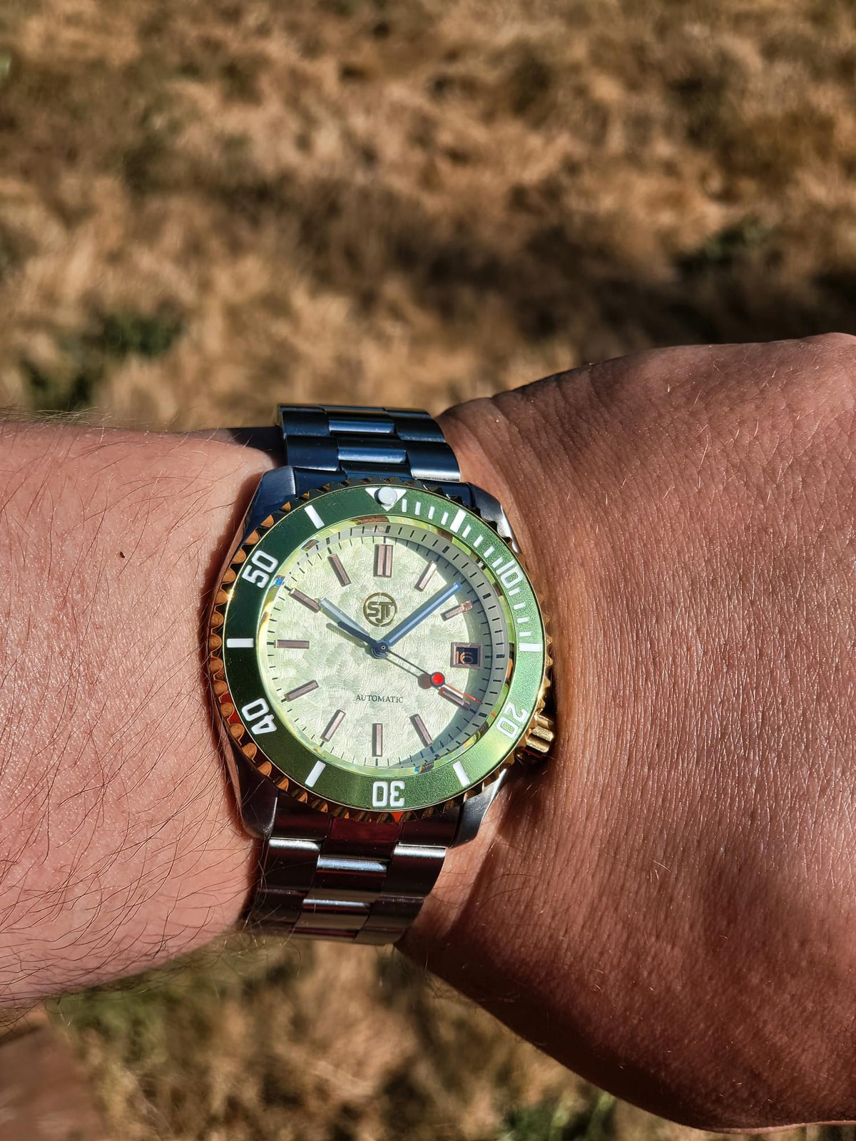 A close-up of a wrist wearing The Coconut SJT Original wristwatch with a green bezel, a cream dial, and a matching green and steel bracelet, set against a blurred outdoor background of dry grass.
