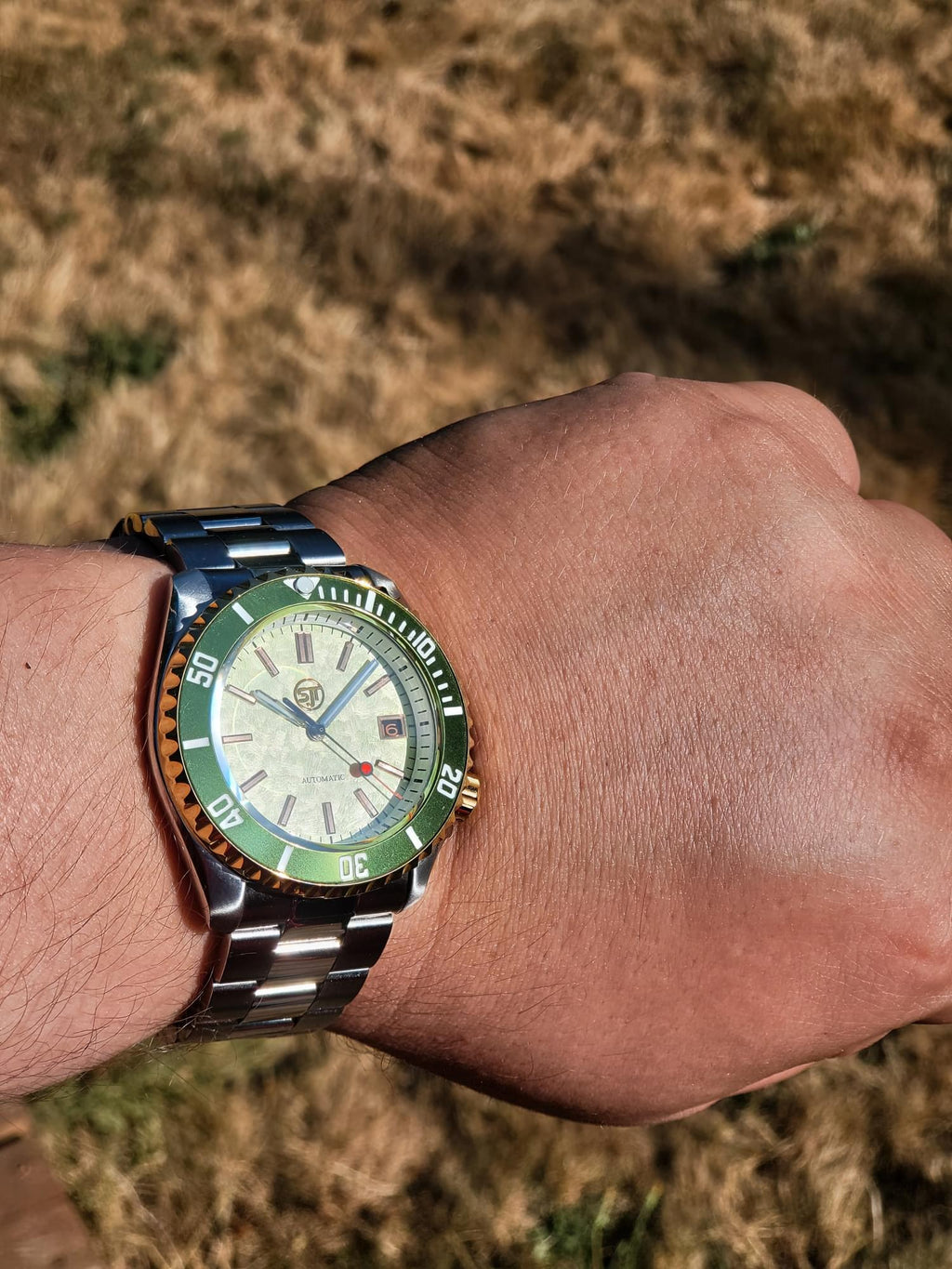 A close-up of a wrist wearing The Coconut SJT Original wristwatch with a green and orange bezel, silver-tone metal bracelet, and a light dial. The background is a blurred outdoor grassy field.