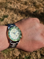 A close-up of a wrist wearing The Coconut SJT Original wristwatch with a green and orange bezel, silver-tone metal bracelet, and a light dial. The background is a blurred outdoor grassy field.