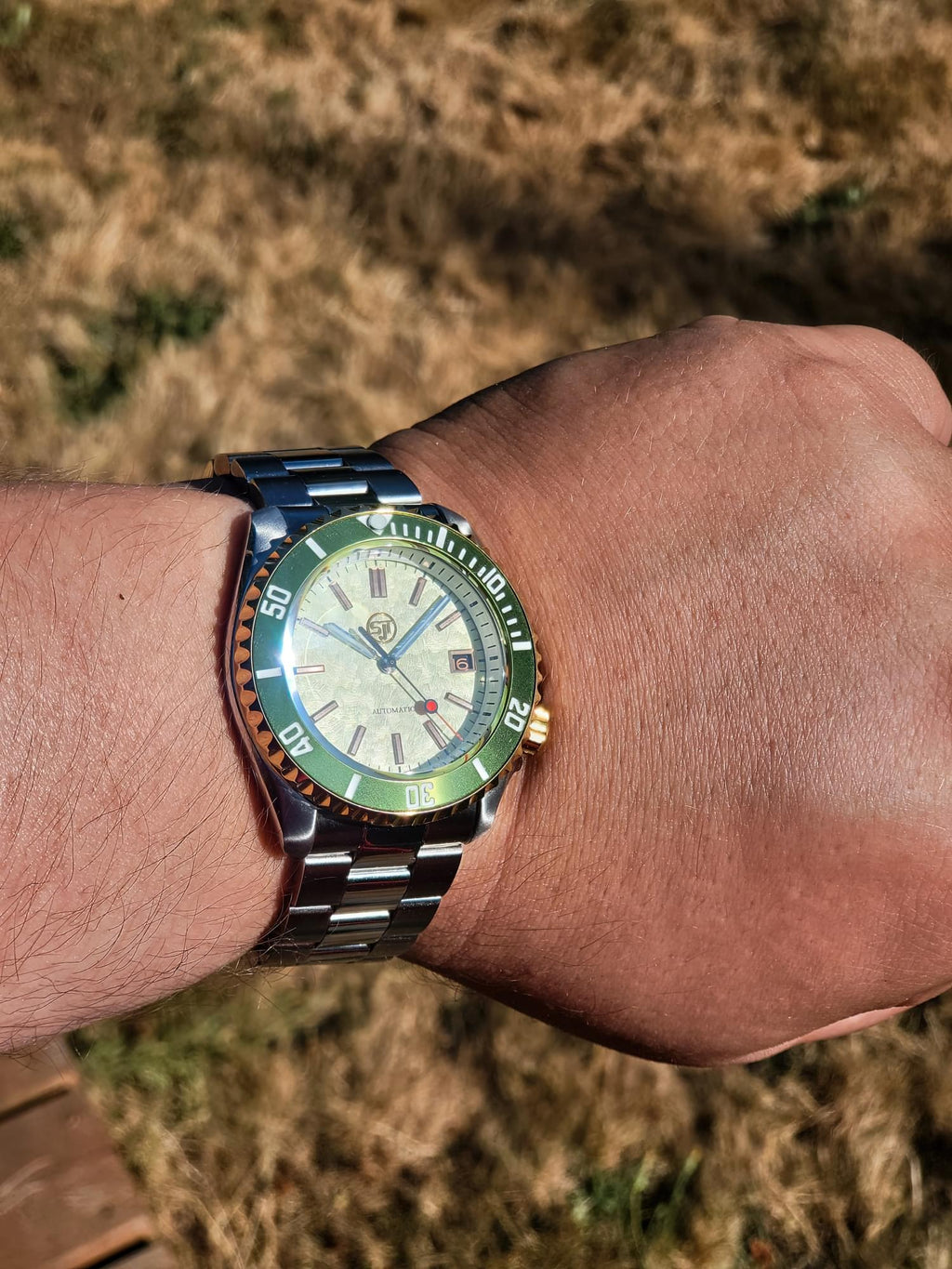 A close-up of a wrist wearing The Coconut SJT Original wristwatch with a green bezel and a cream-colored dial, set against a blurred outdoor background of dry grass.