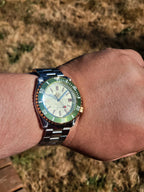 A close-up of a wrist wearing The Coconut SJT Original wristwatch with a green bezel and a cream-colored dial, set against a blurred outdoor background of dry grass.
