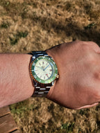 The Coconut SJT Original wristwatch with a green bezel and cream dial, worn on a person's wrist against a dry grass background.
