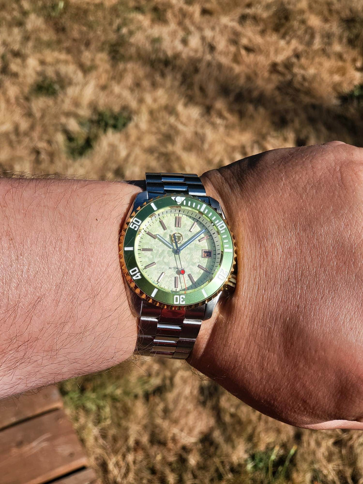 The Coconut SJT Original wristwatch with a green bezel and a light green dial, worn on a person's wrist against a dry grass background.