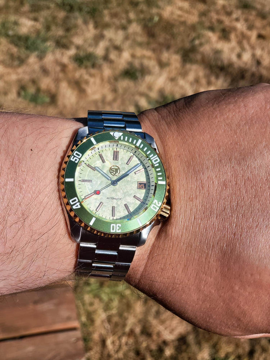 A wrist wearing The Coconut SJT Original wristwatch with a green and orange bezel, a cream-colored dial, and a blue-toned metal bracelet, photographed outdoors against a blurred grassy background.