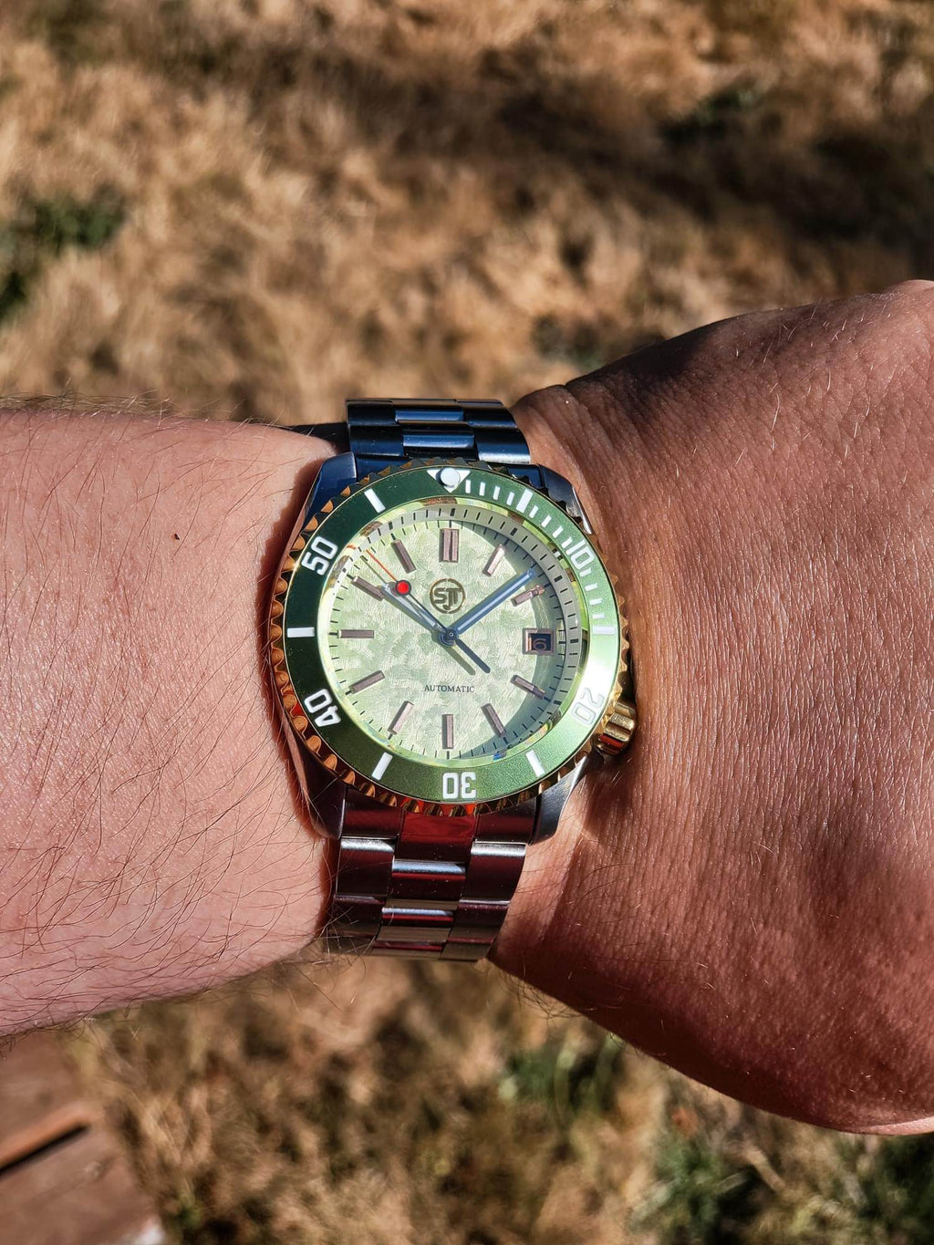 A close-up of a wrist wearing The Coconut SJT Original wristwatch with a green and orange bezel, a light-colored dial, and a blue-toned metal bracelet, photographed outdoors against a blurred dry grass background.