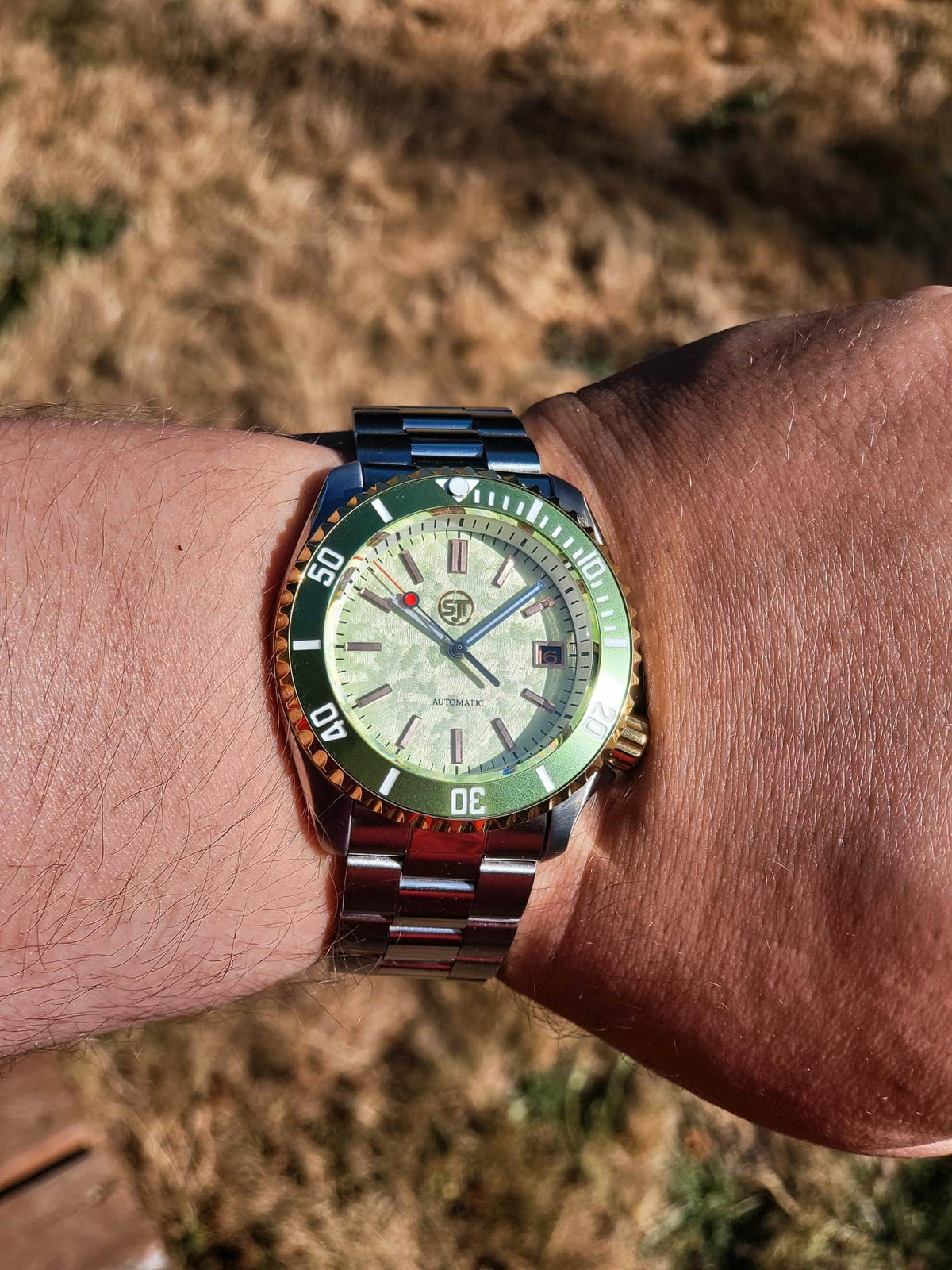 A close-up of a wrist wearing The Coconut SJT Original wristwatch with a green and orange bezel, a light-colored dial, and a blue-toned metal bracelet, photographed outdoors against a blurred dry grass background.