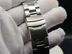 A close-up of The Coconut SJT Original wristwatch band clasp held between white gloved fingers against a dark background.