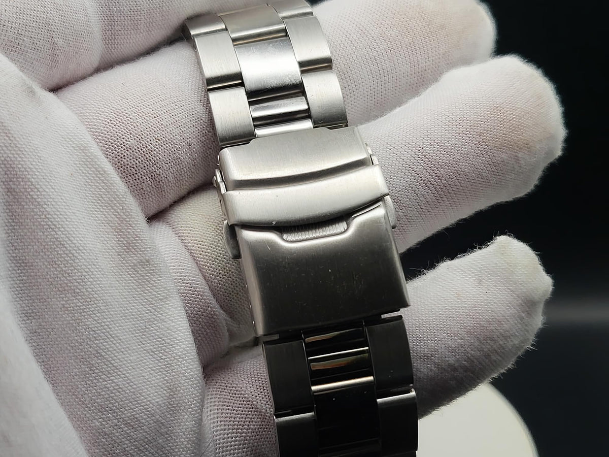 A close-up of The Coconut SJT Original wristwatch band clasp held between white gloved fingers against a dark background.