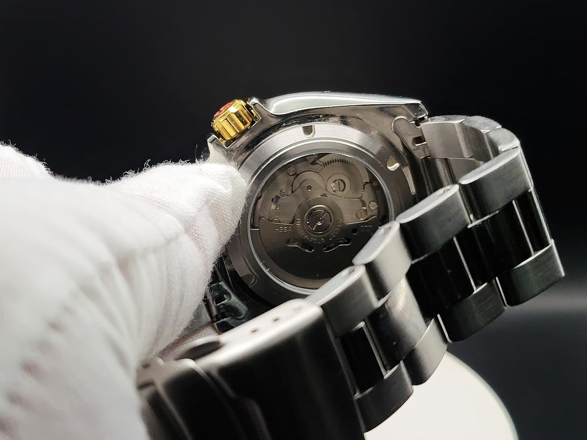 Close-up of a stainless steel automatic watch, The Coconut SJT Original wristwatch, with a transparent case back showing the mechanical movement, held by a white-gloved hand against a dark background.
