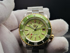 The Coconut SJT Original wristwatch with a green dial and green bezel, held by white-gloved hands against a dark background.