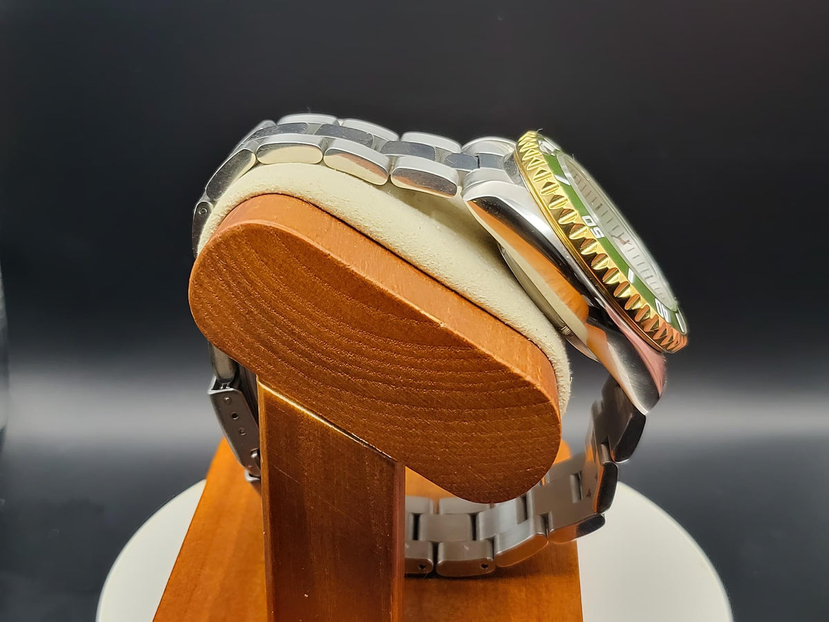 The Coconut SJT Original wristwatch with a gold-tone bezel and green dial accents, displayed on a wooden stand with a cream cushion against a dark background.