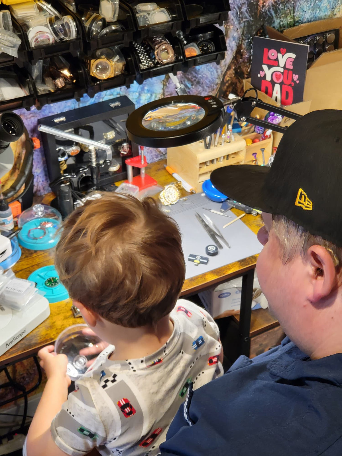 A watchmaker father at work as his child son watches inside a watch repair shop. The man wears a black New Era cap, and the workspace is cluttered with tools, magnifying lamps, and watch parts.
