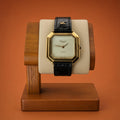 A luxury Léonard Genève watch with an octagonal gold-tone case, cream dial, and black embossed strap, displayed on a wooden stand against a solid terracotta background.