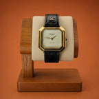 A luxury Léonard Genève watch with an octagonal gold-tone case, cream dial, and black embossed strap, displayed on a wooden stand against a solid terracotta background.
