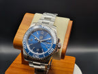 The Memo SJT Original wristwatch with a blue dial and blue bezel, displayed on a wooden stand with a beige cushion against a dark background.