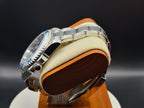 The Memo SJT Original wristwatch with a blue bezel and silver metal bracelet, displayed on a cream cushion with a brown wooden base against a dark background.
