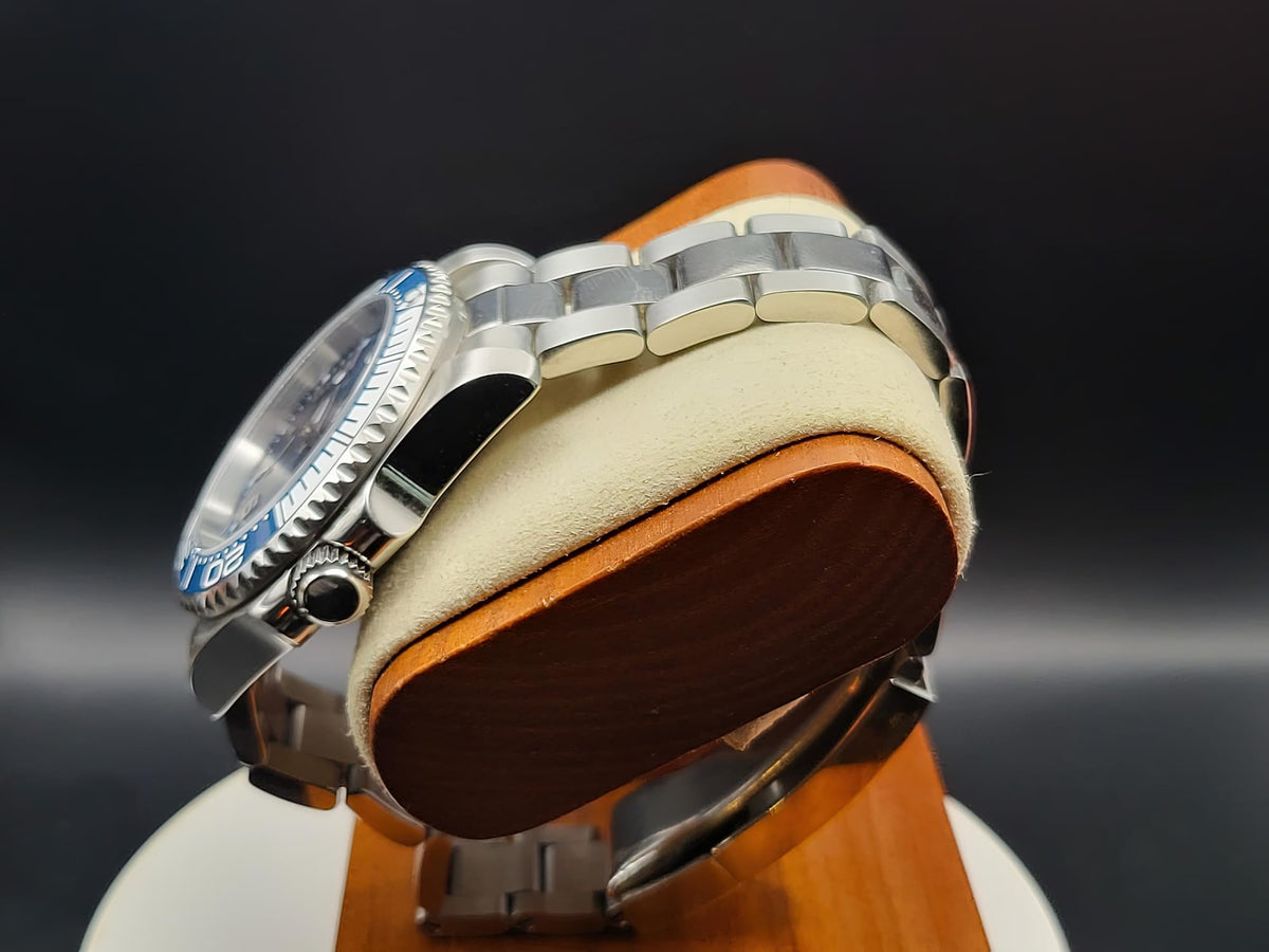 The Memo SJT Original wristwatch with a blue bezel and silver metal bracelet, displayed on a cream cushion with a brown wooden base against a dark background.