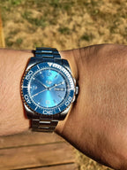 The Memo SJT Original wristwatch with a blue dial and blue bezel, worn on a person's wrist outdoors with a blurred tan grassy background.
