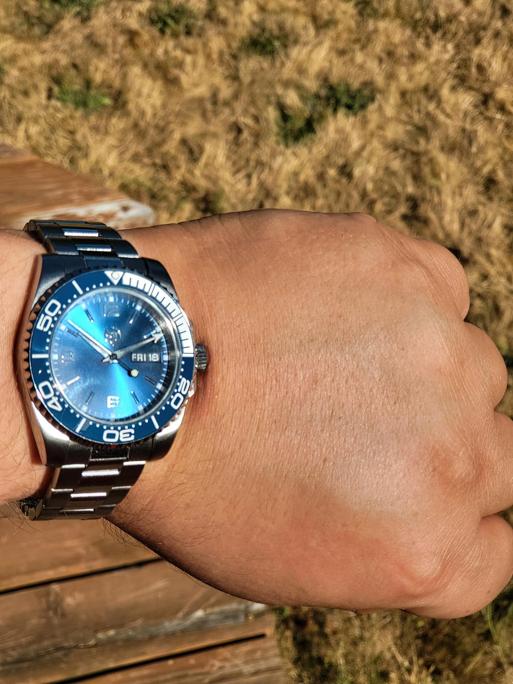 The Memo SJT Original wristwatch with a blue dial and blue bezel, worn on a person's wrist against a background of dry grass and a wooden deck.