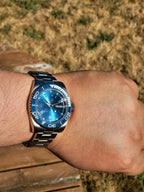 The Memo SJT Original wristwatch with a blue dial and blue bezel, worn on a person's wrist against a blurred outdoor background of dry grass and wooden boards.