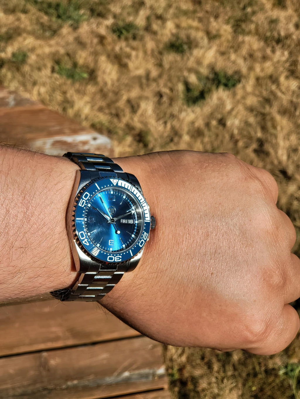 A close-up of a wrist wearing The Memo SJT Original wristwatch with a vibrant blue sunburst dial and blue bezel, set against a blurred background of dry grass and a wooden surface.