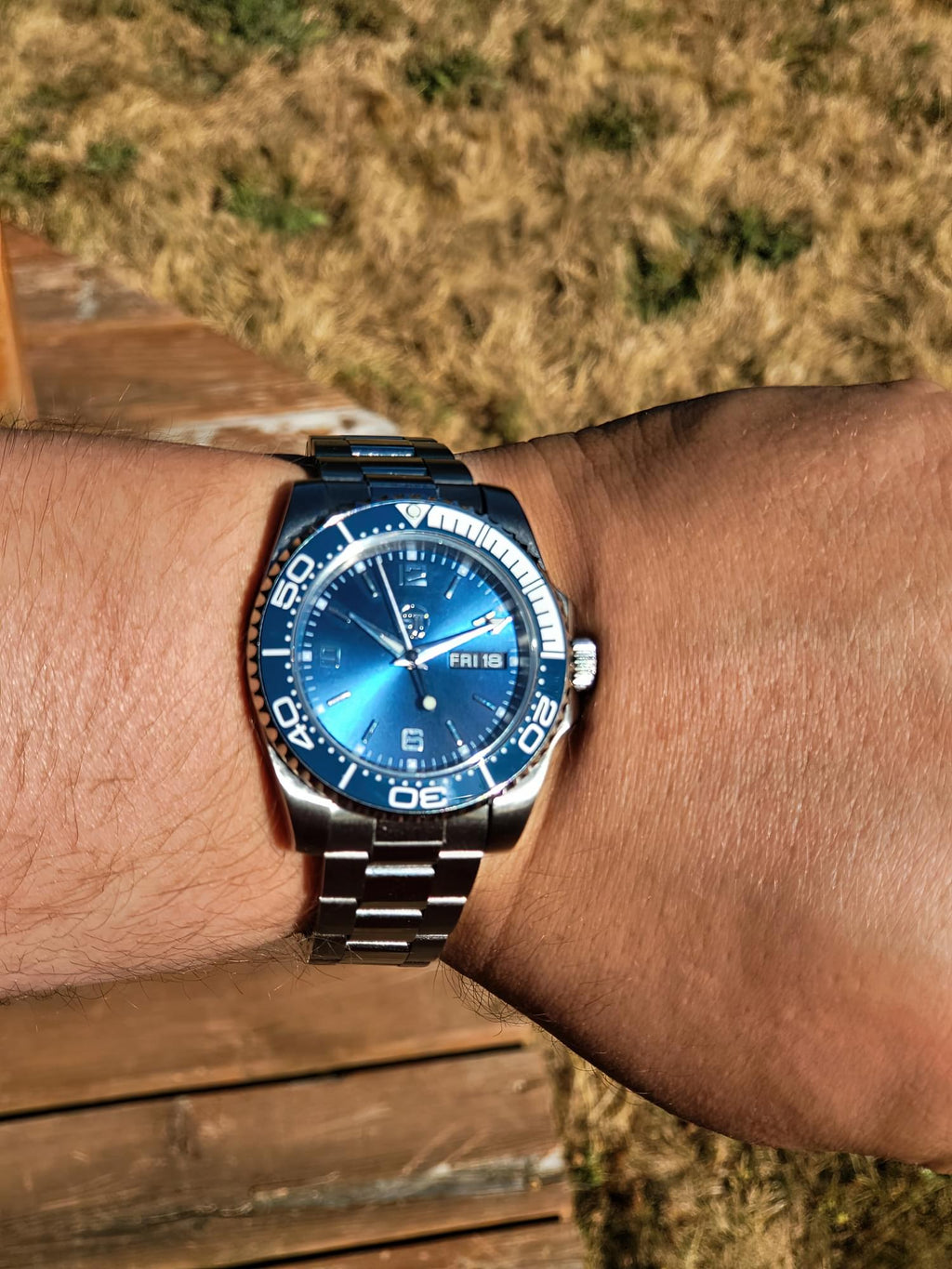 The Memo SJT Original wristwatch with a blue sunburst dial and blue bezel, worn on a person's wrist outdoors on a wooden deck with dry grass in the background.