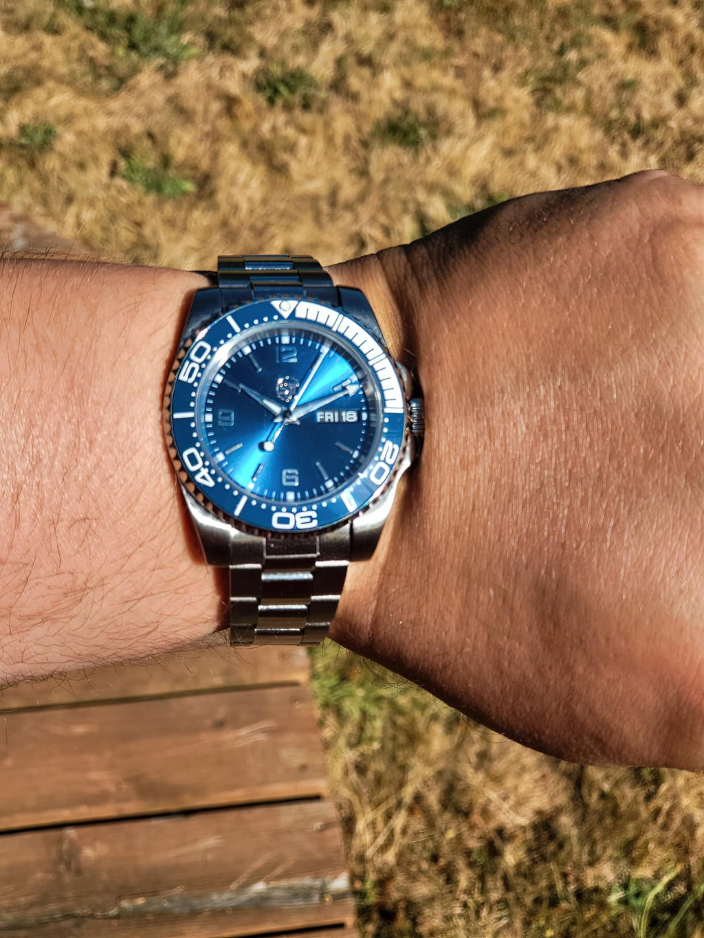 The Memo SJT Original wristwatch with a blue sunburst dial and a silver stainless steel bracelet, shown worn on a person's wrist outdoors with dry grass and a wooden deck in the background.