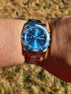 A wrist wearing The Memo SJT Original wristwatch with a matching blue and silver link bracelet, photographed outdoors against a blurred dry grass background.