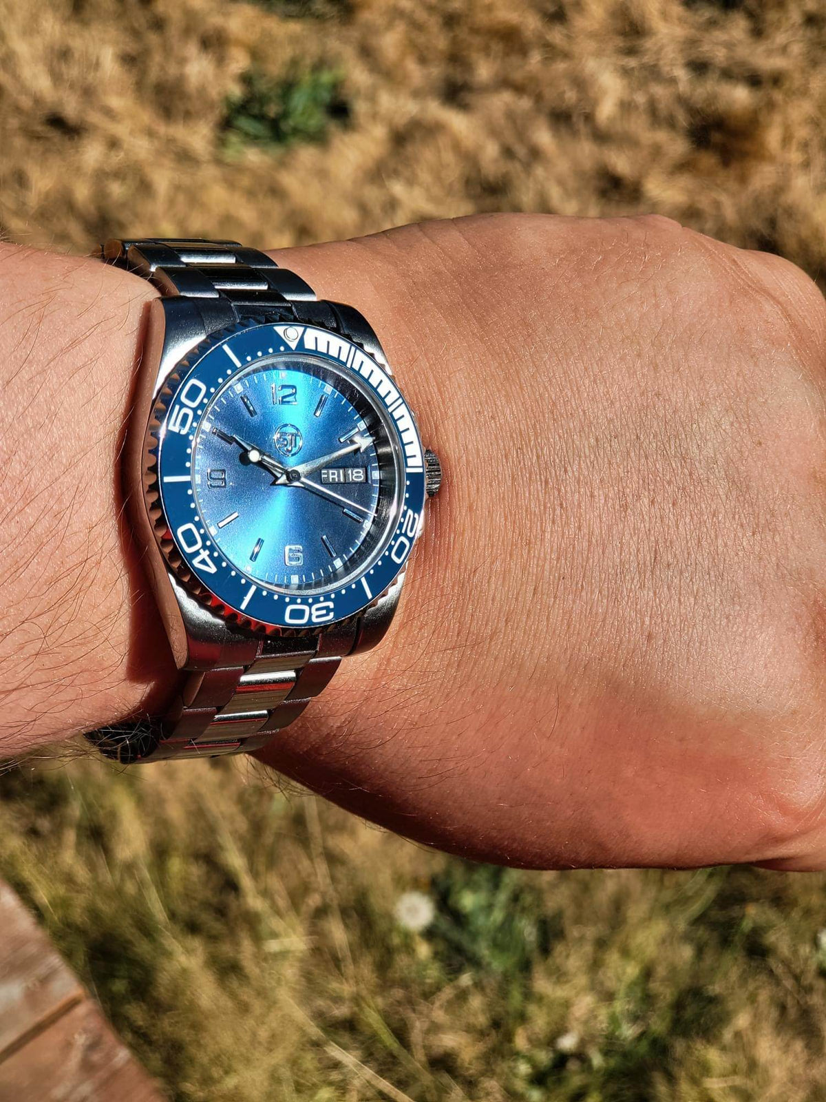 A wrist wearing The Memo SJT Original wristwatch with a blue dial and blue bezel, photographed outdoors against a blurred grassy background.