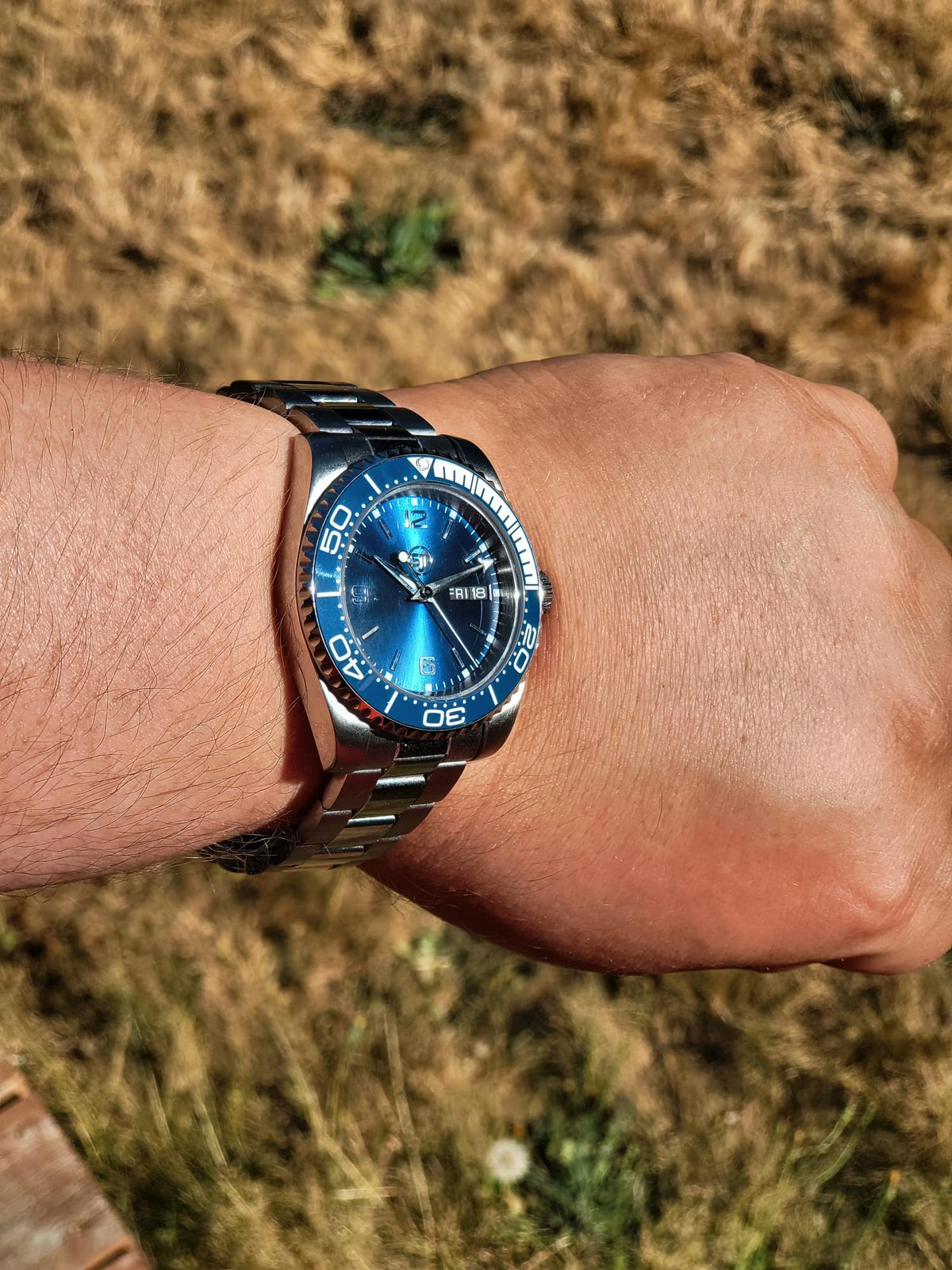 A wrist wearing The Memo SJT Original wristwatch with a vibrant blue dial and blue bezel, photographed outdoors against a blurred background of dry grass.