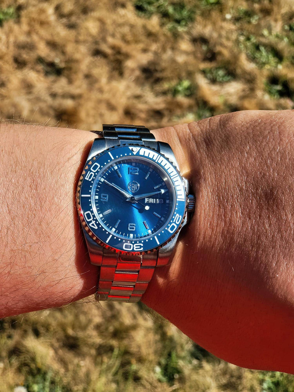 The Memo SJT Original wristwatch with a blue dial and blue-and-red metal bracelet, worn on a person's wrist outdoors with dry grass in the background.