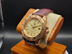 The Midas SJT Original wristwatch with a rose gold-toned case, textured champagne dial, and a dark reddish-brown leather strap, displayed on a wooden stand.