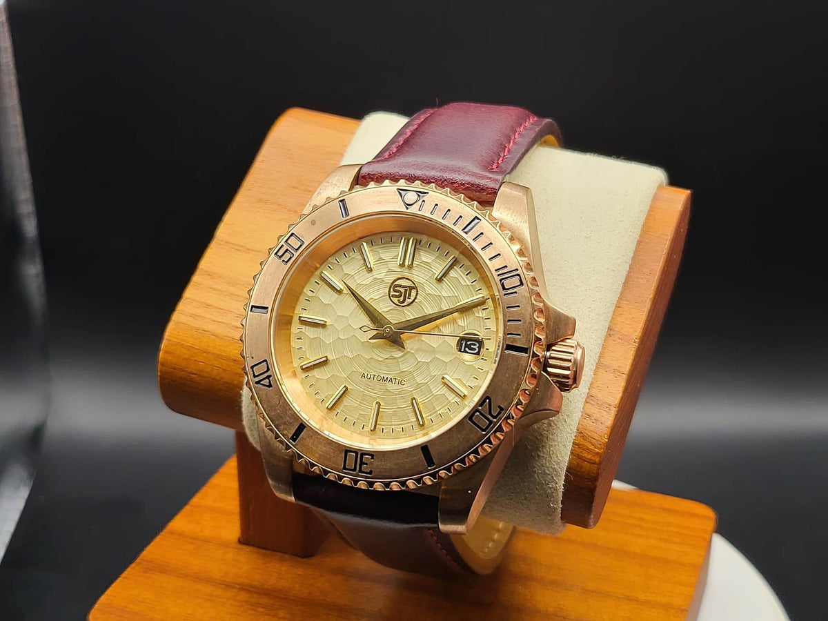 The Midas SJT Original wristwatch with a rose gold-toned case, textured champagne dial, and a burgundy leather strap, displayed on a wooden stand against a dark background.