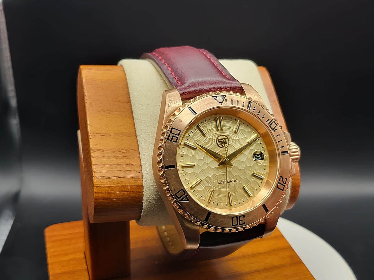 The Midas SJT Original wristwatch with a rose gold-toned case, gold dial, and burgundy leather strap, displayed on a wooden stand against a dark background.