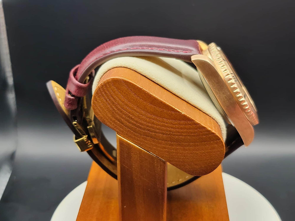 A close-up of The Midas SJT Original wristwatch with a burgundy leather strap, gold-toned case, and cream-colored cushion on a wooden display stand.