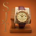 The Midas SJT Original wristwatch with a rose gold-toned case, textured champagne dial, and dark burgundy leather strap, displayed on a wooden watch stand against a warm orange background with a gold 'SJT' logo on the left.
