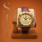 The Midas SJT Original wristwatch with a rose gold-toned case, textured champagne dial, and dark burgundy leather strap, displayed on a wooden watch stand against a warm orange background with a gold 'SJT' logo on the left.