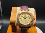 The Midas SJT Original wristwatch with a rose gold-toned case, textured champagne dial, and a burgundy leather strap, displayed on a wooden stand against a dark background.