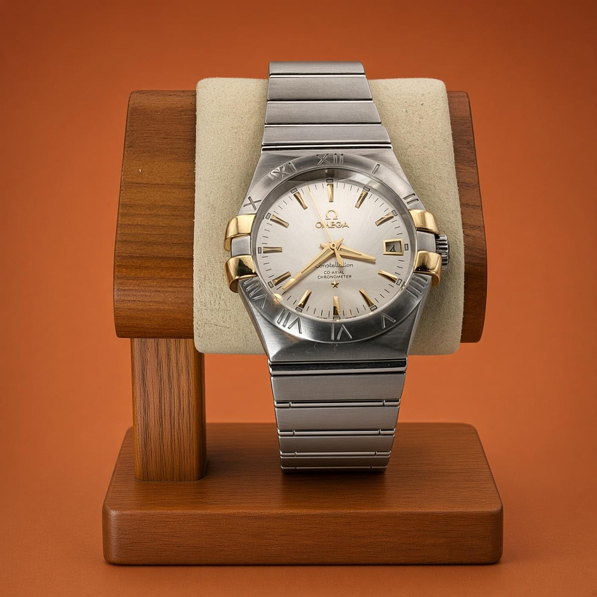 An Omega Constellation wristwatch with a two-tone silver stainless steel and gold-tone case, featuring a white dial and silver link bracelet, displayed on a wooden stand against a solid orange background.