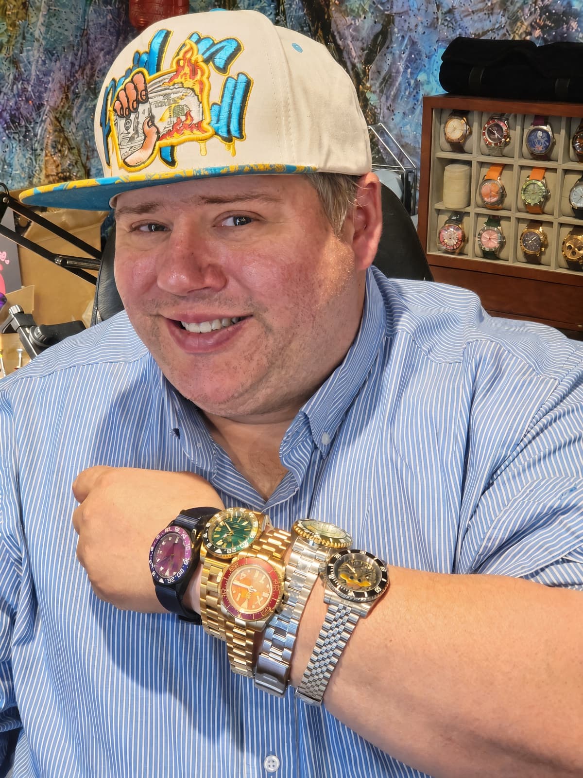 A smiling man wearing a light blue striped button-down shirt and a beige baseball cap with a colorful graphic patch, showcasing a stack of five different luxury wristwatches on his left wrist in an indoor setting.