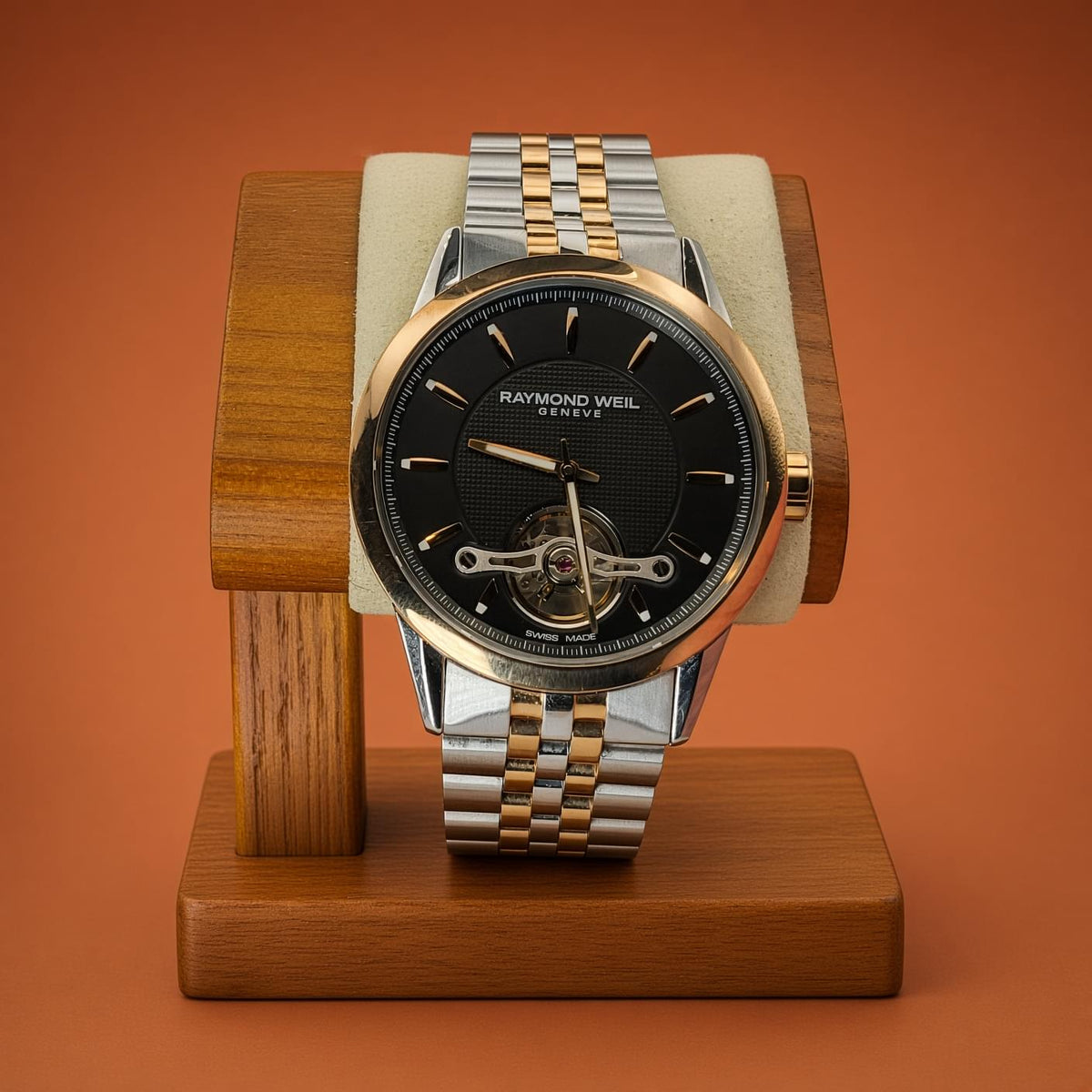 A Raymond Weil watch with a two-tone silver and gold stainless steel bracelet, displayed on a wooden stand with a cream cushion against a solid terracotta background.
