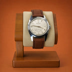 A vintage-style Reliance wristwatch with a silver-toned case, cream dial, and brown leather strap, displayed upright on a wooden watch stand against a warm orange-brown background.