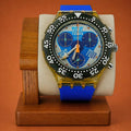 A Swatch wristwatch with a blue strap, gold-toned case, black bezel, and a blue dial featuring the words 'TWELVE' 'FOUR' and 'EIGHT', displayed on a wooden stand against a warm orange-brown background.