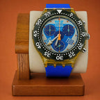 A Swatch wristwatch with a blue strap, gold-toned case, black bezel, and a blue dial featuring the words 'TWELVE' 'FOUR' and 'EIGHT', displayed on a wooden stand against a warm orange-brown background.