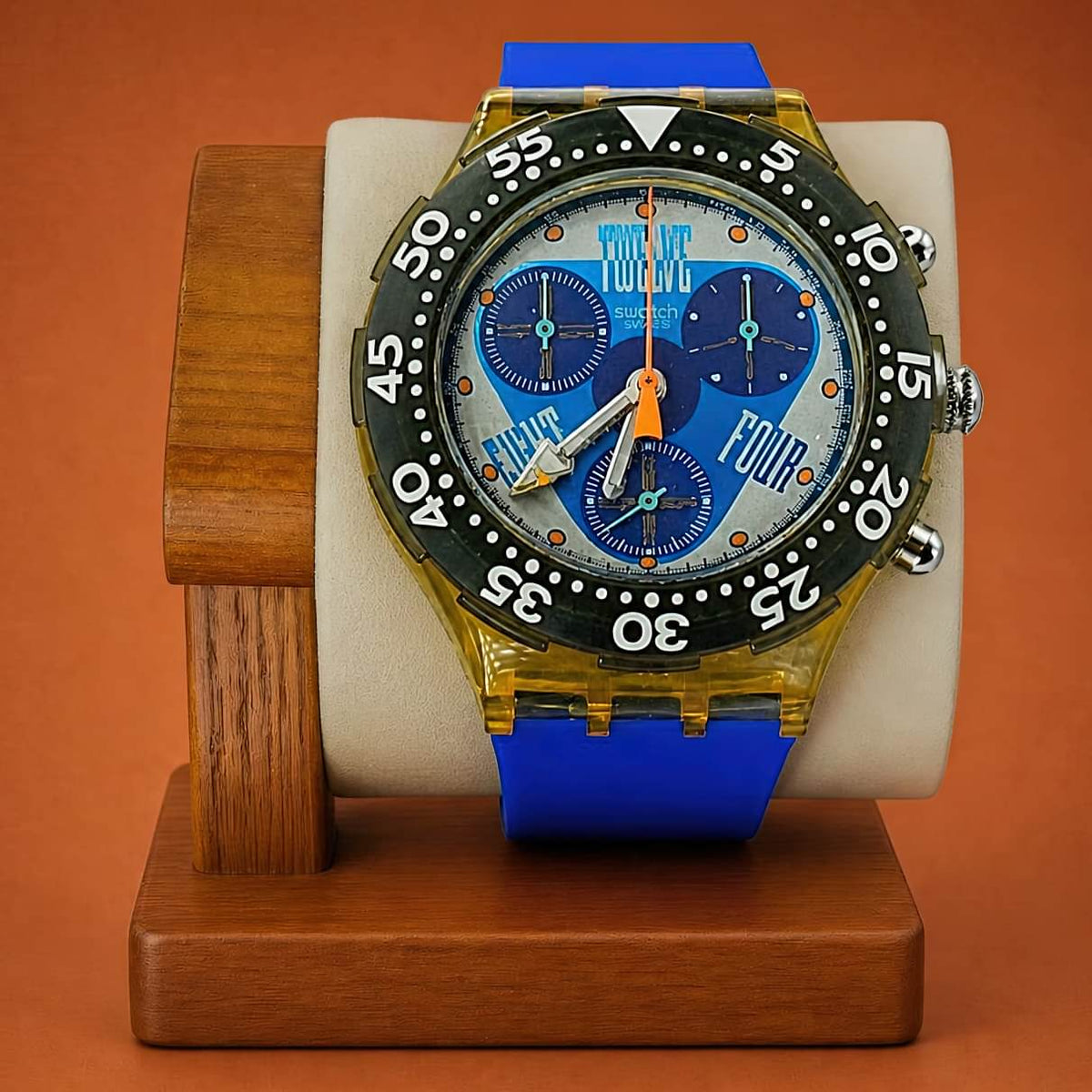 A Swatch wristwatch with a blue strap, gold-toned case, black bezel, and a blue dial featuring the words 'TWELVE' 'FOUR' and 'EIGHT', displayed on a wooden stand against a warm orange-brown background.