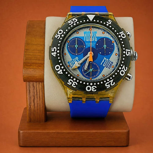 A Swatch wristwatch with a blue strap, gold-toned case, black bezel, and a blue dial featuring the words 'TWELVE' 'FOUR' and 'EIGHT', displayed on a wooden stand against a warm orange-brown background.