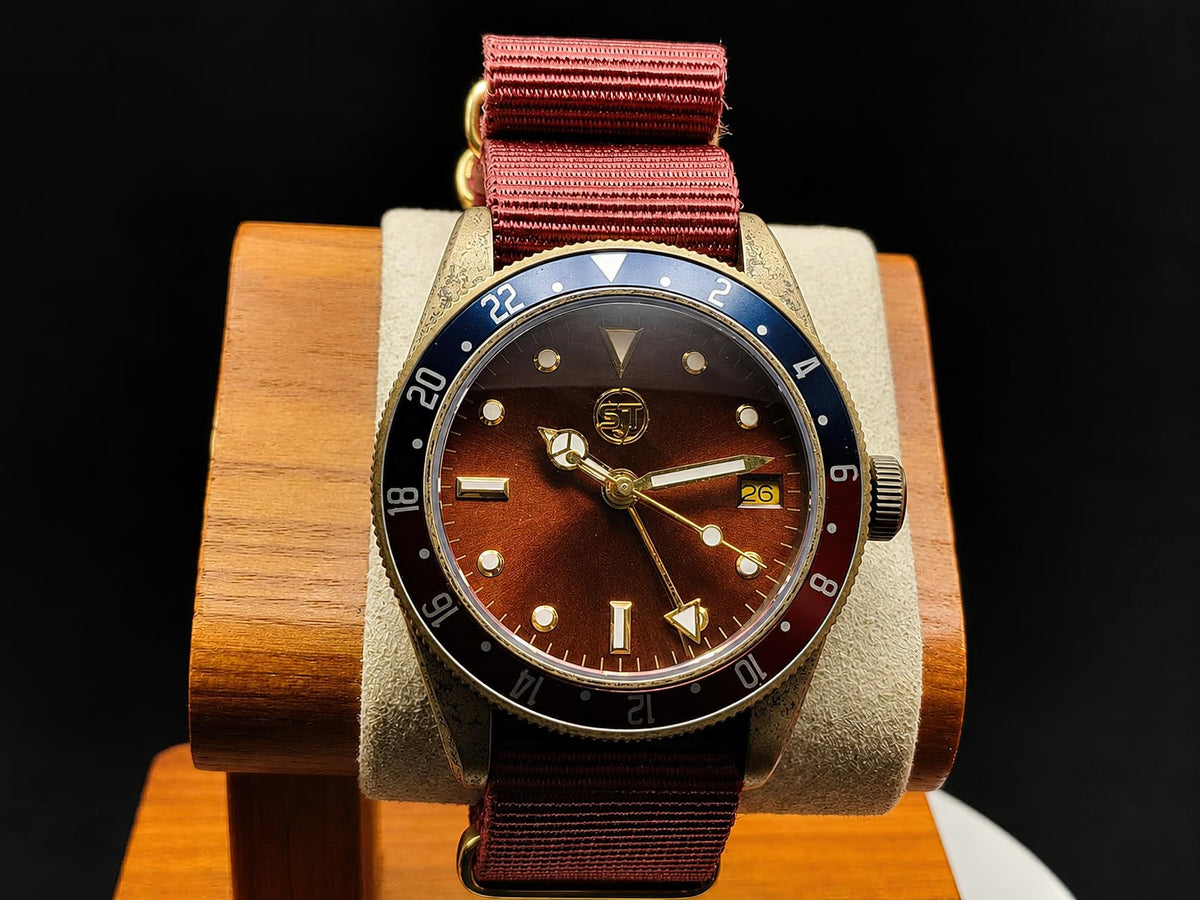 The Vintner SJT Original wristwatch with a brown sunburst dial, a blue and red bezel, and a reddish-brown fabric strap, displayed on a wooden stand against a black background.