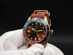 The Vintner SJT Original wristwatch with a gold-toned case, brown dial, red and blue bezel, and red fabric strap, shown resting on a white gloved hand against a black background.